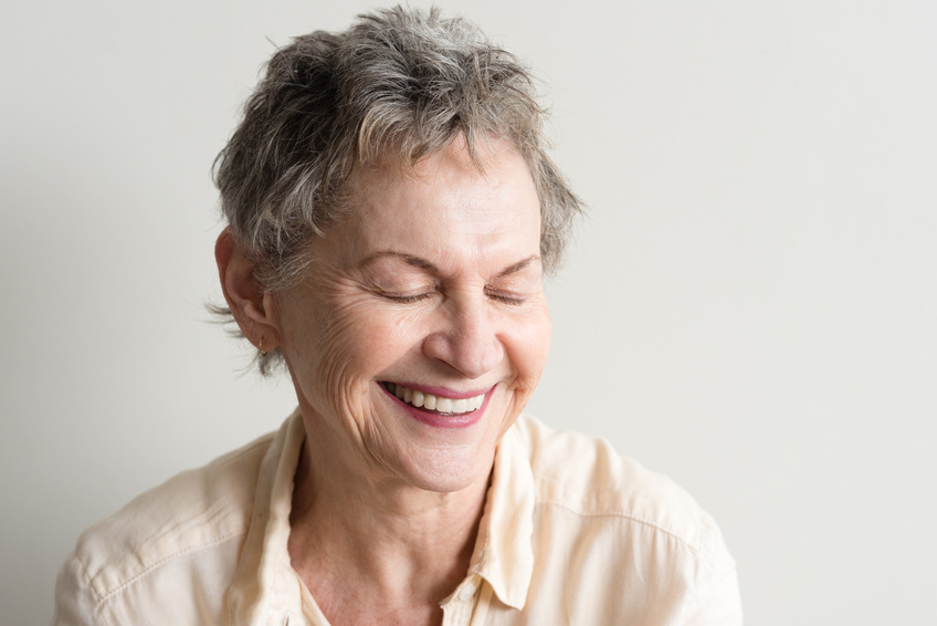 Head and shoulders view of older woman with short grey hair laughing with eyes closed against neutral background (selective focus)