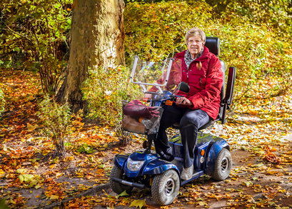 Seniorin fährt mit ihrem Elektromobil durch einen herbstlichen Wald spazieren und lächelt in die Kamera