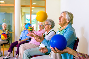 Group of happy senior ladies doing coordination exercises in a seniors gym sitting in chairs throwing and catching brightly colored balls.