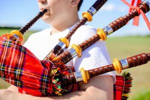 Closeup picture of young man playing traditional Scotland bagpipe on green summer outdoors copy space background