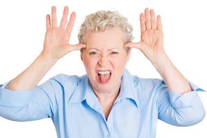 Closeup portrait of senior mature woman sticking out tongue at you, camera gesture, thumbs hands on temple, isolated white background. Negative human emotions, facial expressions, feelings, attitude