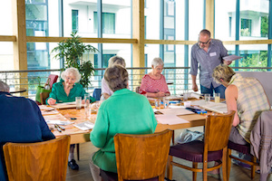 Male art instructor wearing eyeglasses overseeing group of cute senior students painting with watercolor.
