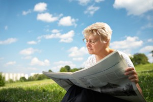 The woman in old age sits on a grass in park on a background of the blue sky and reads the newspaper